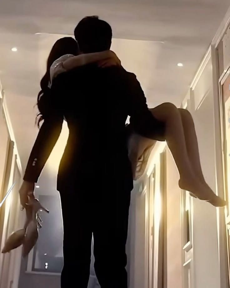 A man carries a woman down a softly lit hallway at night, her high heels dangling from her hand