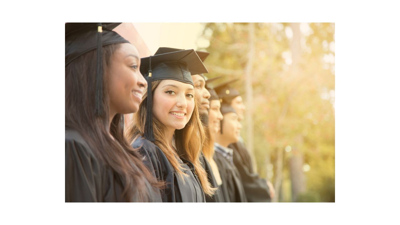 A group of college students at graduation day are standing in their caps and gowns, in profile, while one student is turned toward the camera and smiling.