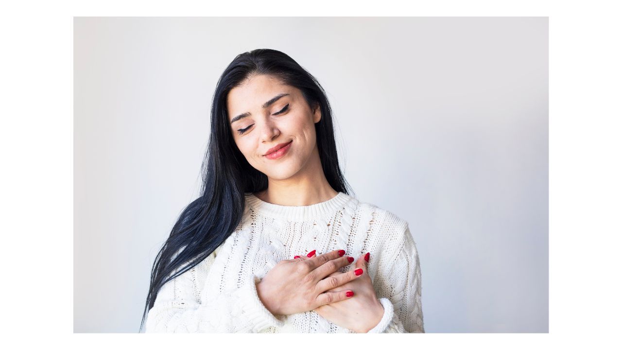 A young person is standing in front of a plain, white wall, smiling with her eyes closed and both hands held up to her heart, like she is giving herself a hug.