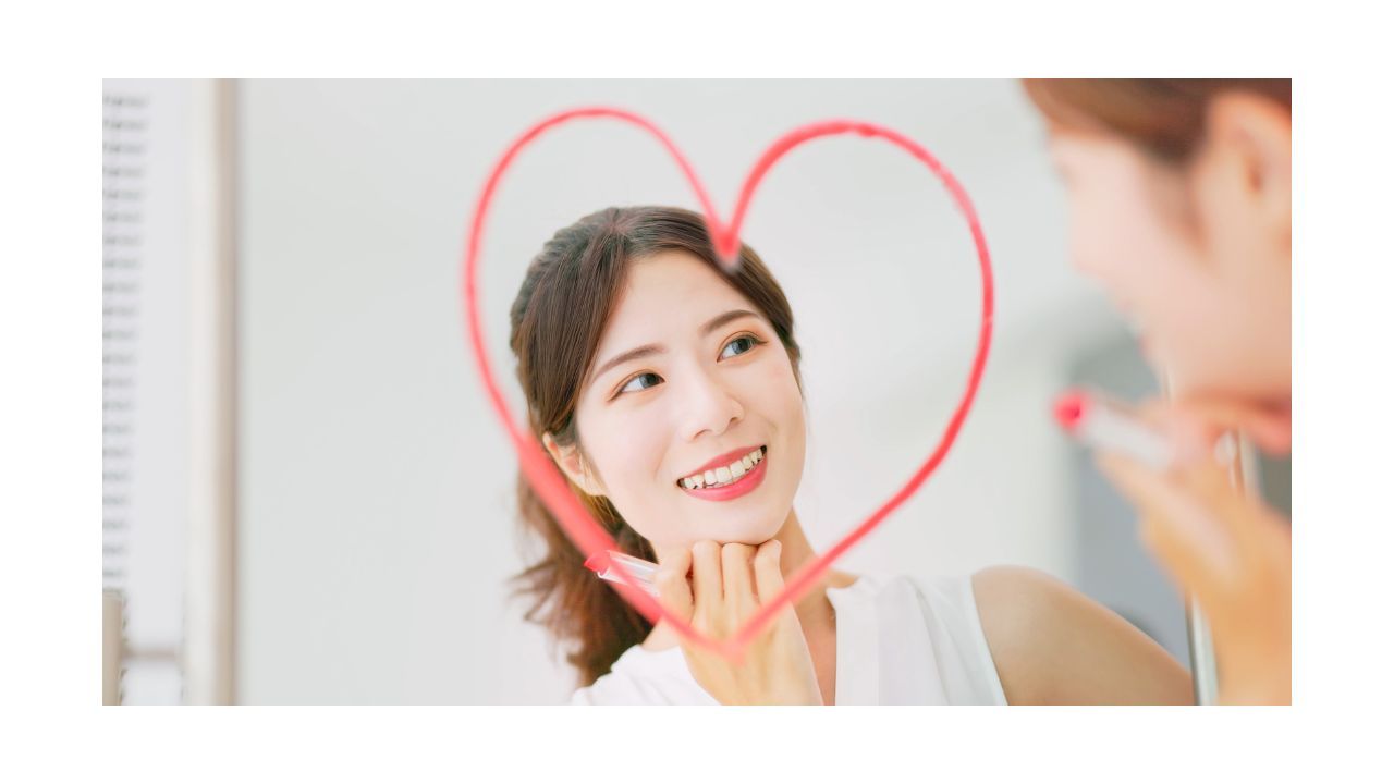 A young person is looking at her reflection in a mirror and smiling. She is holding a tube of coral-colored lipstick and has drawn a colorful heart around her face in the mirror. She is happy and joyful.