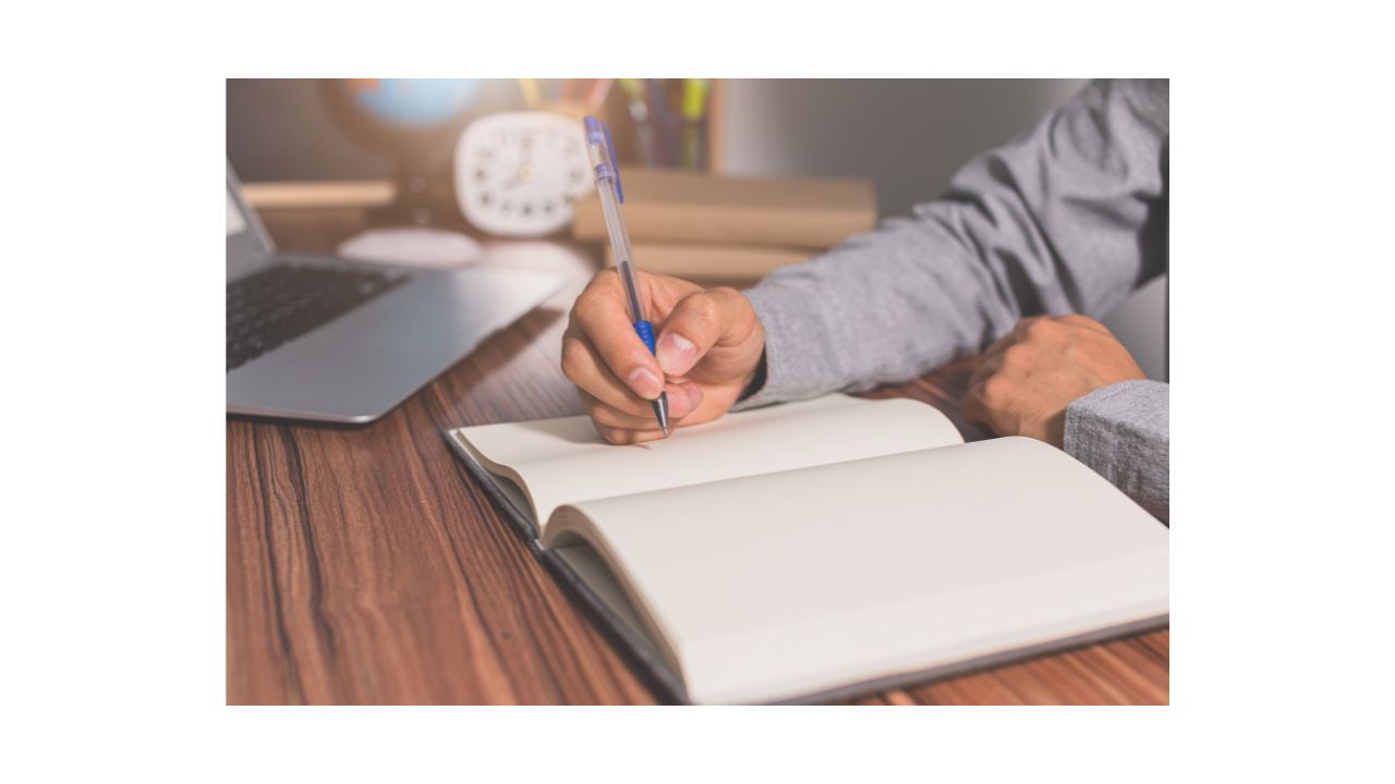 A person is sitting at a desk in front of an open notebook, holding a pen and appearing to write a letter.