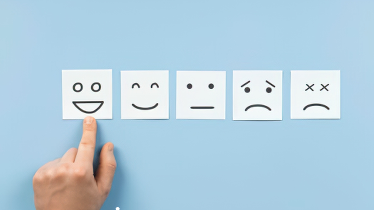 A line of post-its each with a different emoji such has laughing, happy, sad, and neutral are posted onto a pale blue background with a someone's finger pointing to the happy expression post-it. 