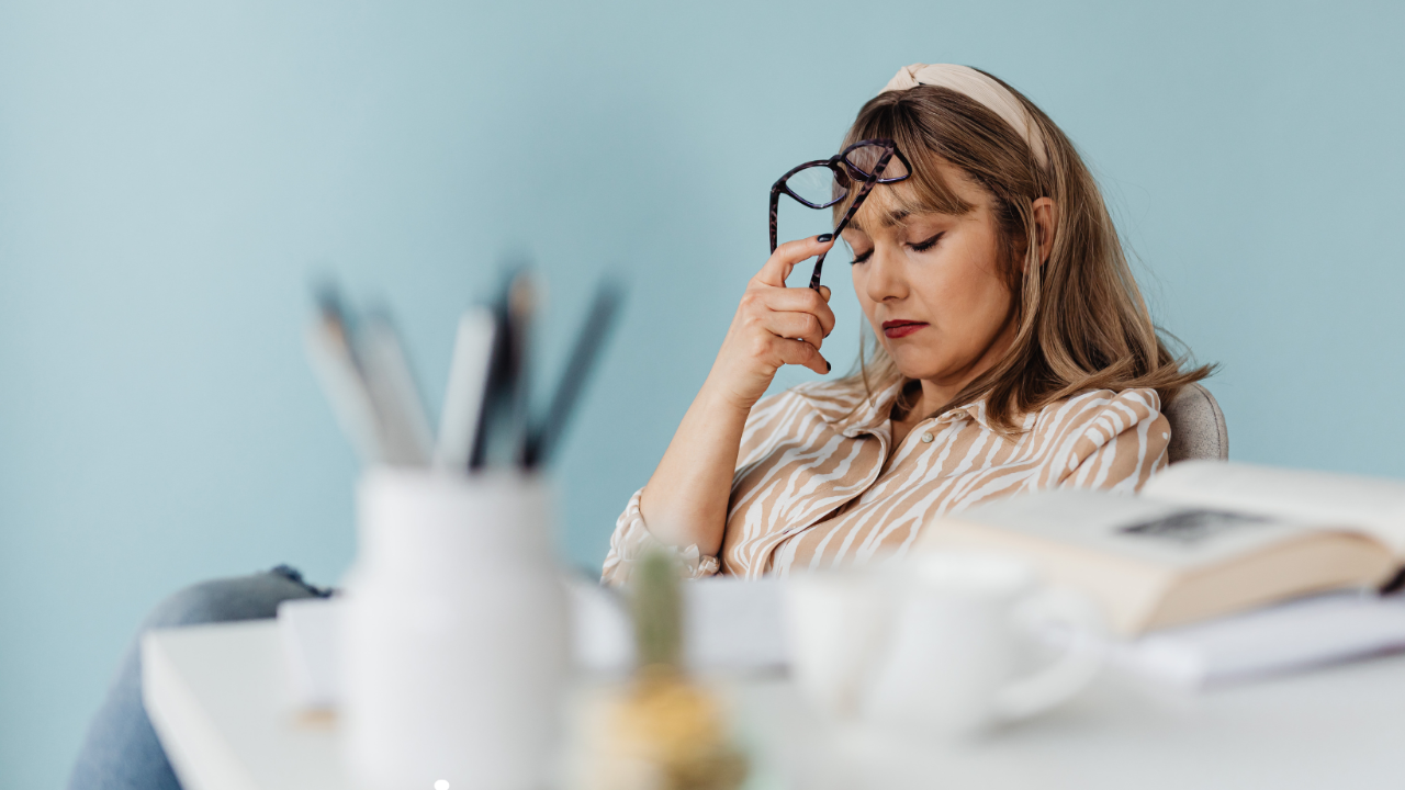 A business woman sits at her desk with a pair of black glasses pressed against her forehead as if she's stressed or overwhelmed.