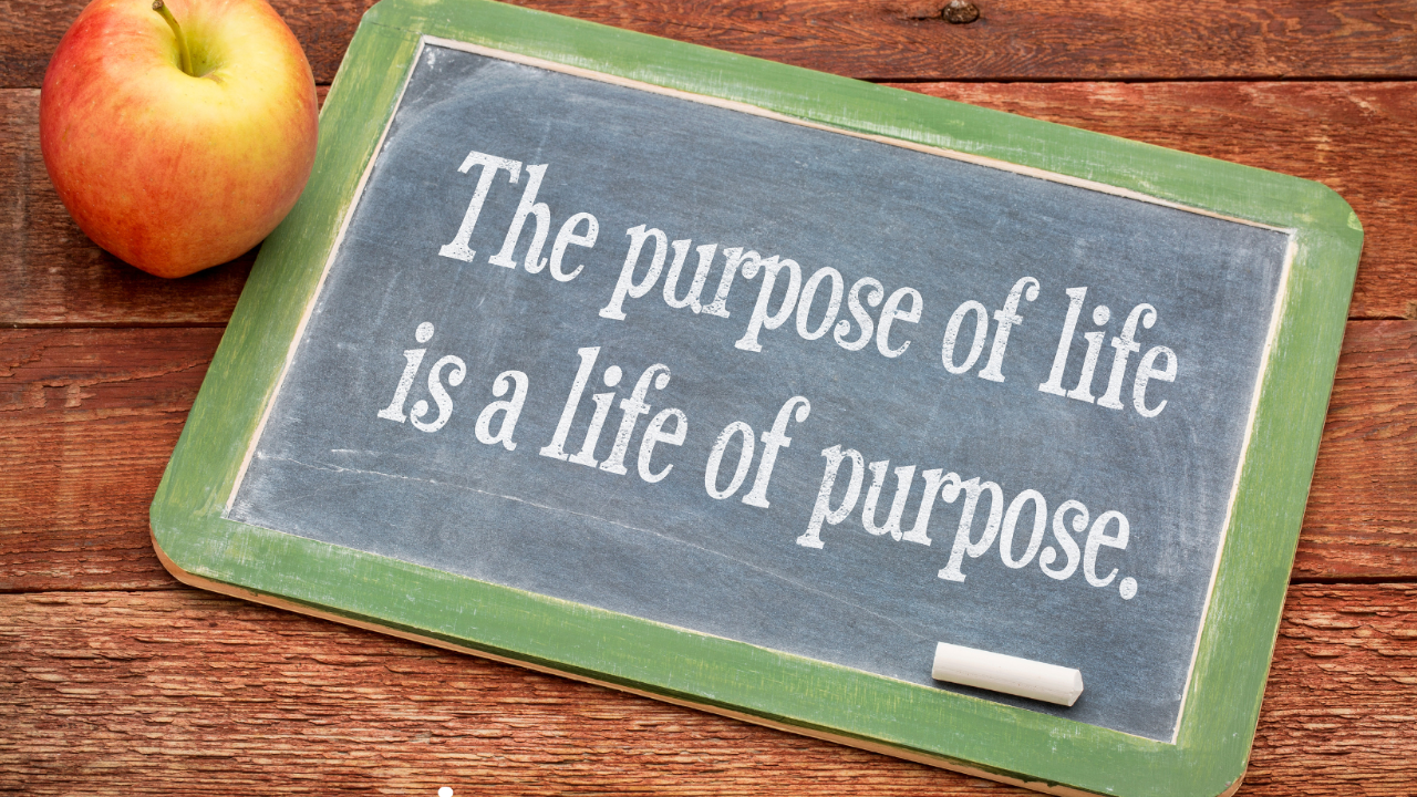 A small chalkboard with a green frame sits on a brown wooden desk with an apple next to the chalkboard. The chalkboard has the phrase "The purpose of life is a life of purpose".