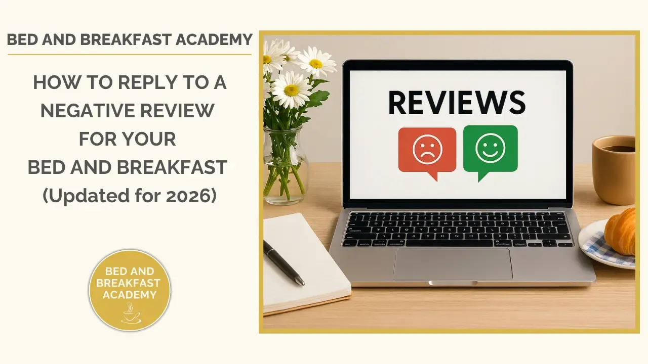 A laptop on a wooden desk displays the word “REVIEWS” with unhappy and happy icons. Beside it sit a cup of coffee, a plate with a croissant on a blue checked napkin, a notebook with a pen, and a vase of white daisies. The left side of the image shows the Bed and Breakfast Academy title and the blog heading “How to Reply to a Negative Review for Your Bed and Breakfast (Updated for 2026)”.