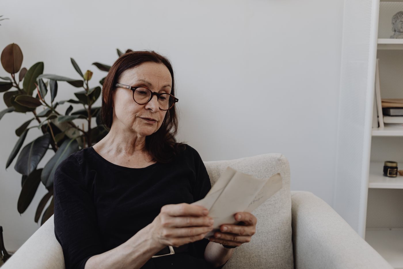 Woman in round glasses sitting calmly in a cream armchair beside a rubber plant, reading an opened letter held in both hands, facing one piece of post instead of hiding it in a drawer