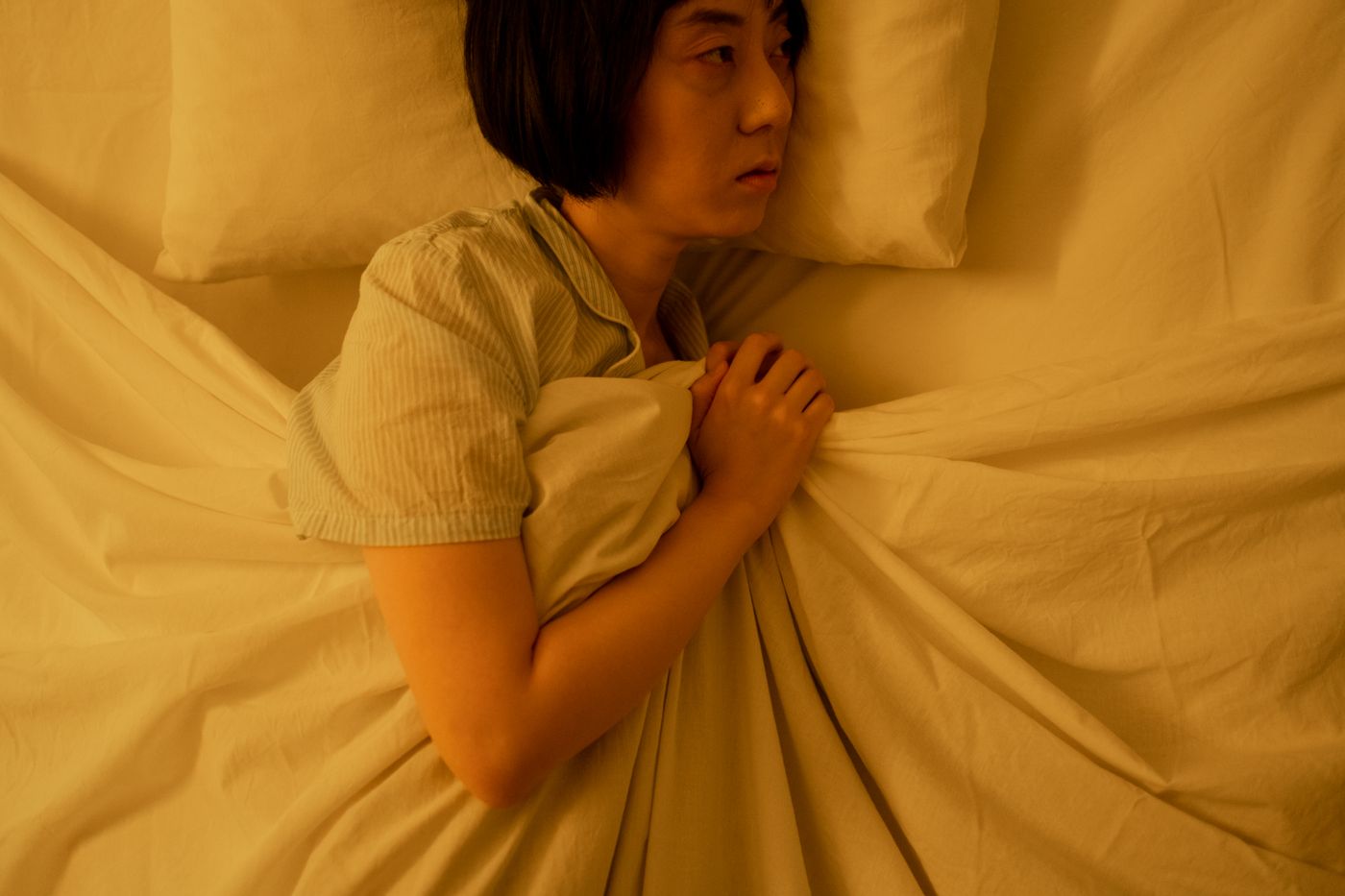 Woman with a dark bob lying awake in bed in warm lamplight, clutching the white duvet to her chest and staring towards the edge of the bed, the wide awake replay of an awkward conversation that keeps running long after the lights go off