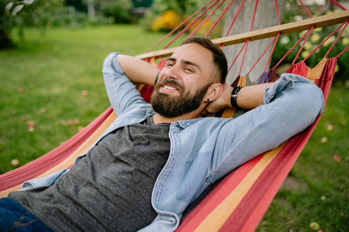 Smiling bearded man lying back in a striped hammock in a garden with his hands behind his head and windfall apples on the grass, taking ten minutes to do nothing while the jobs wait