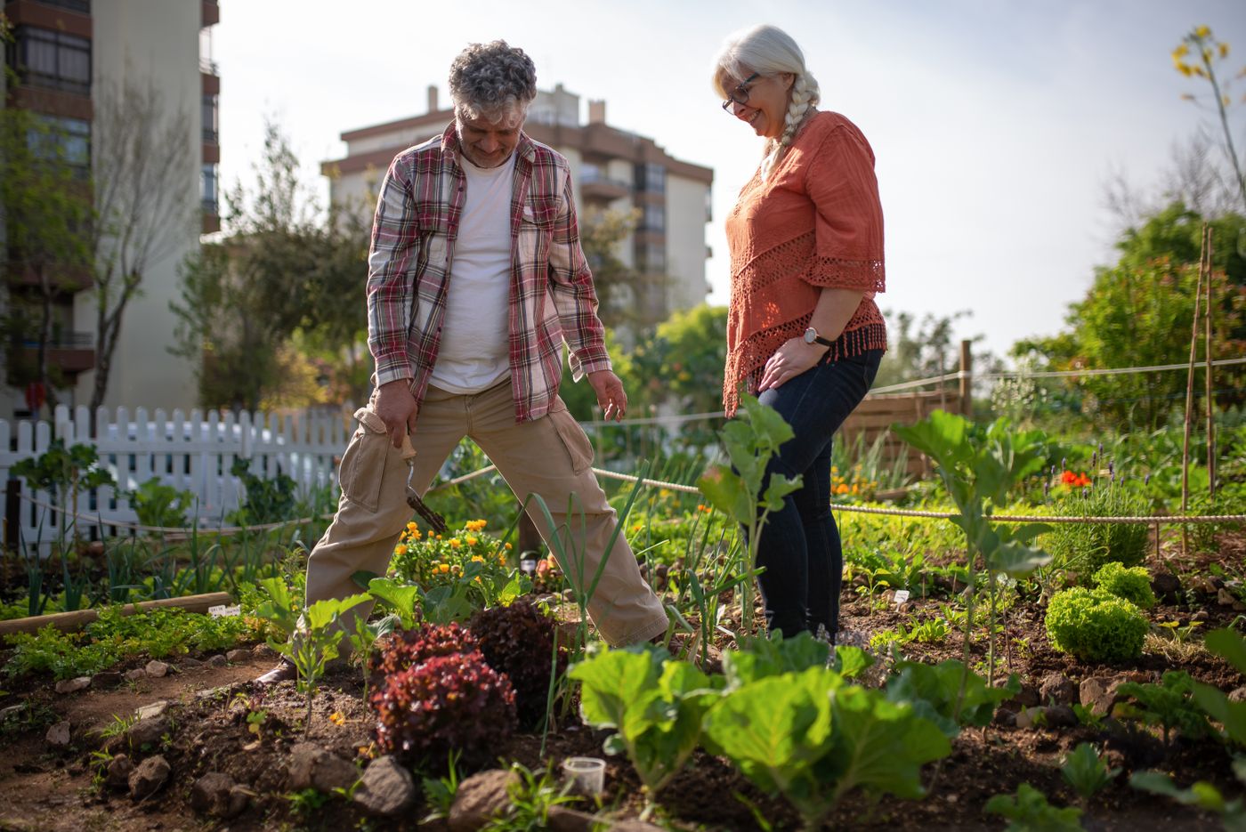 Smiling older couple standing in a sunlit community vegetable garden, the man leaning on a hoe among young lettuces and marigolds, an hour given to planting something that will outlast the afternoon instead of chasing the next milestone