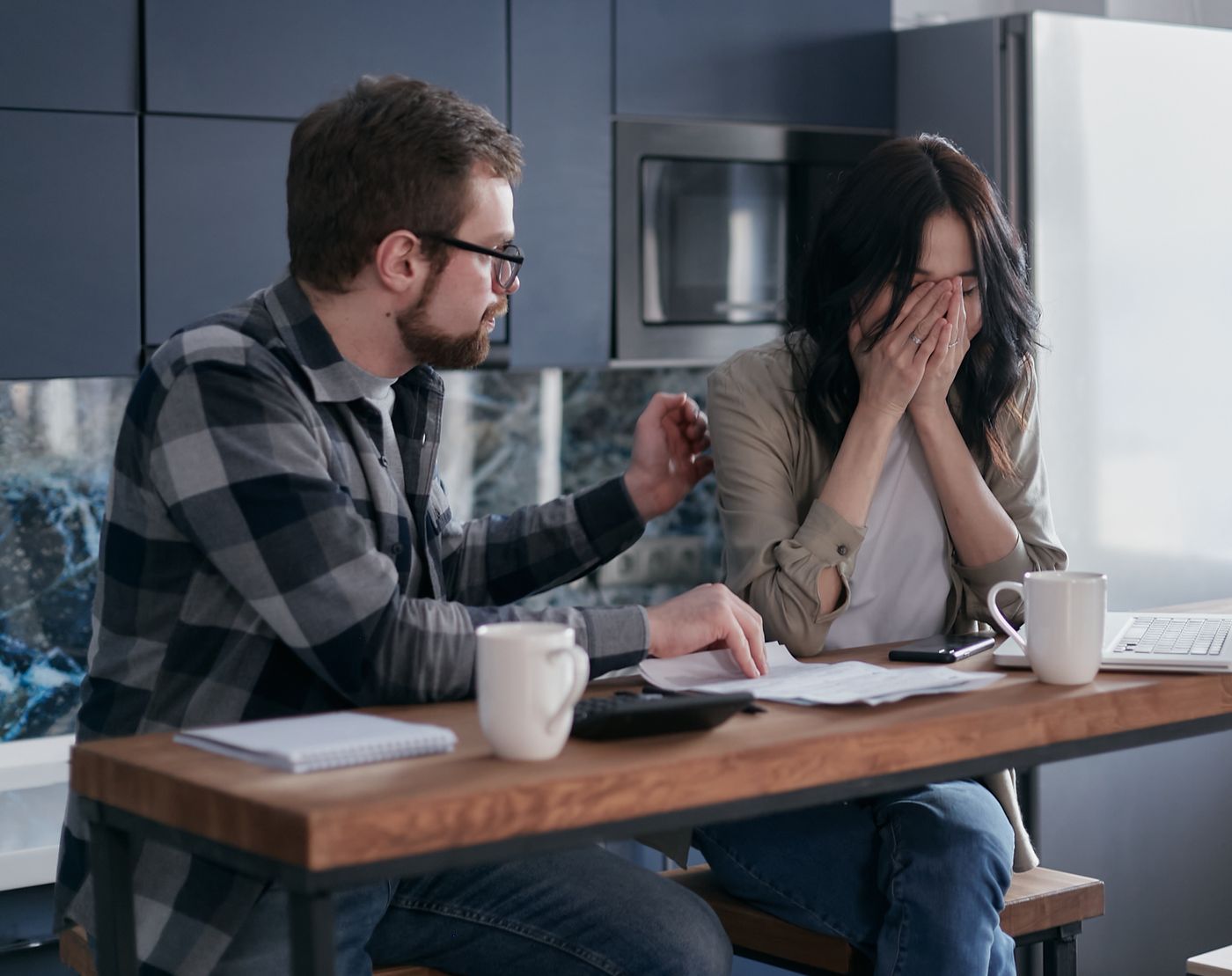 Woman at a kitchen table covering her face with both hands while her partner points at a printed bill beside a calculator and coffee mugs, the flinch that makes not looking at money feel safer than looking