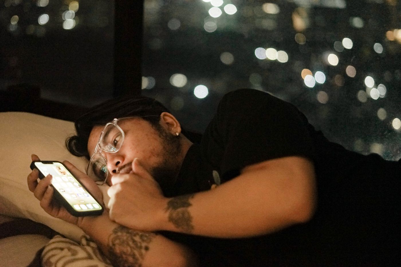 Man with glasses lying in bed at night, scrolling his phone by its glow with city lights blurred through the window behind him, the late Sunday evening scroll that tries to stretch the weekend and quiet the dread of Monday
