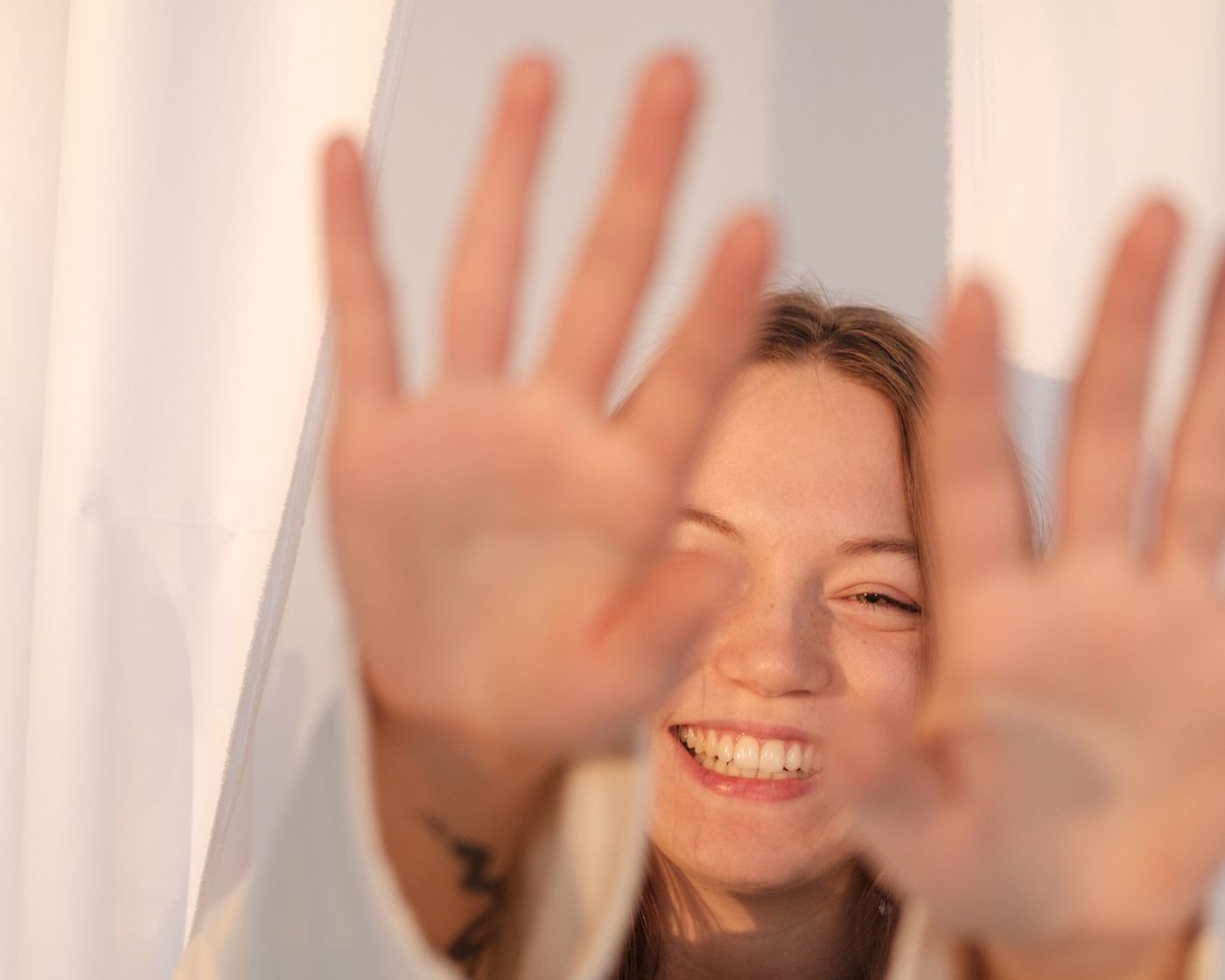 Smiling young woman in a cream jumper holding both palms up towards the camera to hide her face, the same reflex that bats a compliment away before it can land