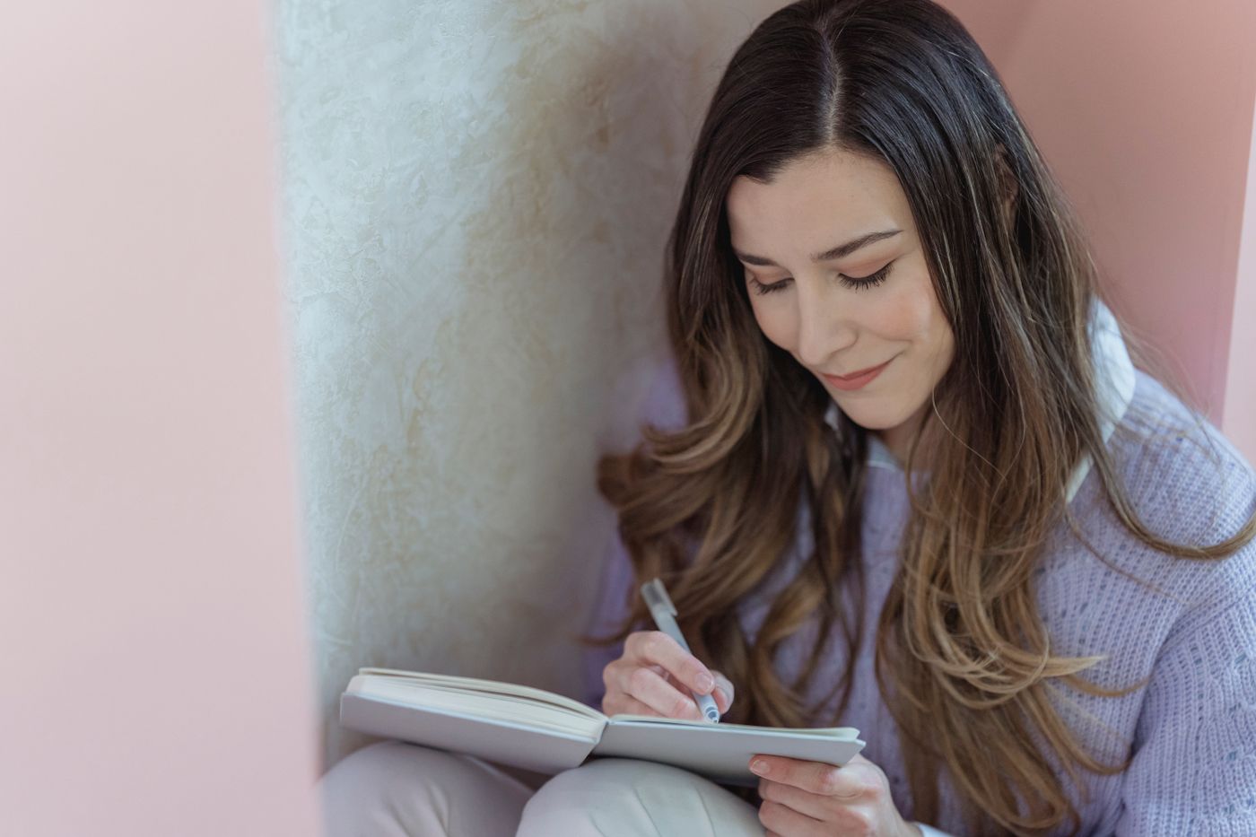 Smiling woman with long brown hair sitting in a quiet corner and writing in a small notebook, turning a pang of comparison into a short note about what she wants for herself
