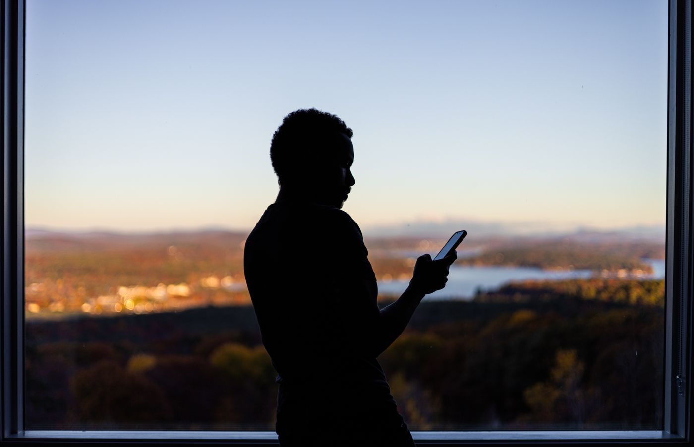 Silhouette of a person standing at a tall window at dusk, looking down at the glowing screen of a smartphone in one hand, with autumn hills, a lake and distant town lights behind them, the quiet moment you reach for the phone to call someone who has gone