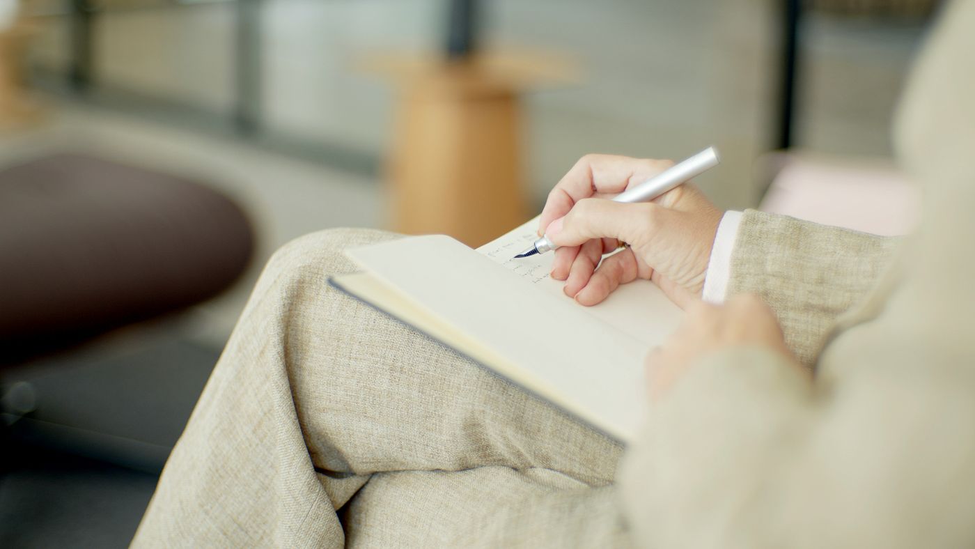 Person in a linen suit writing with a silver pen in a notebook resting on their lap, capturing the inner critic's exact words so negative self-talk can be examined on paper