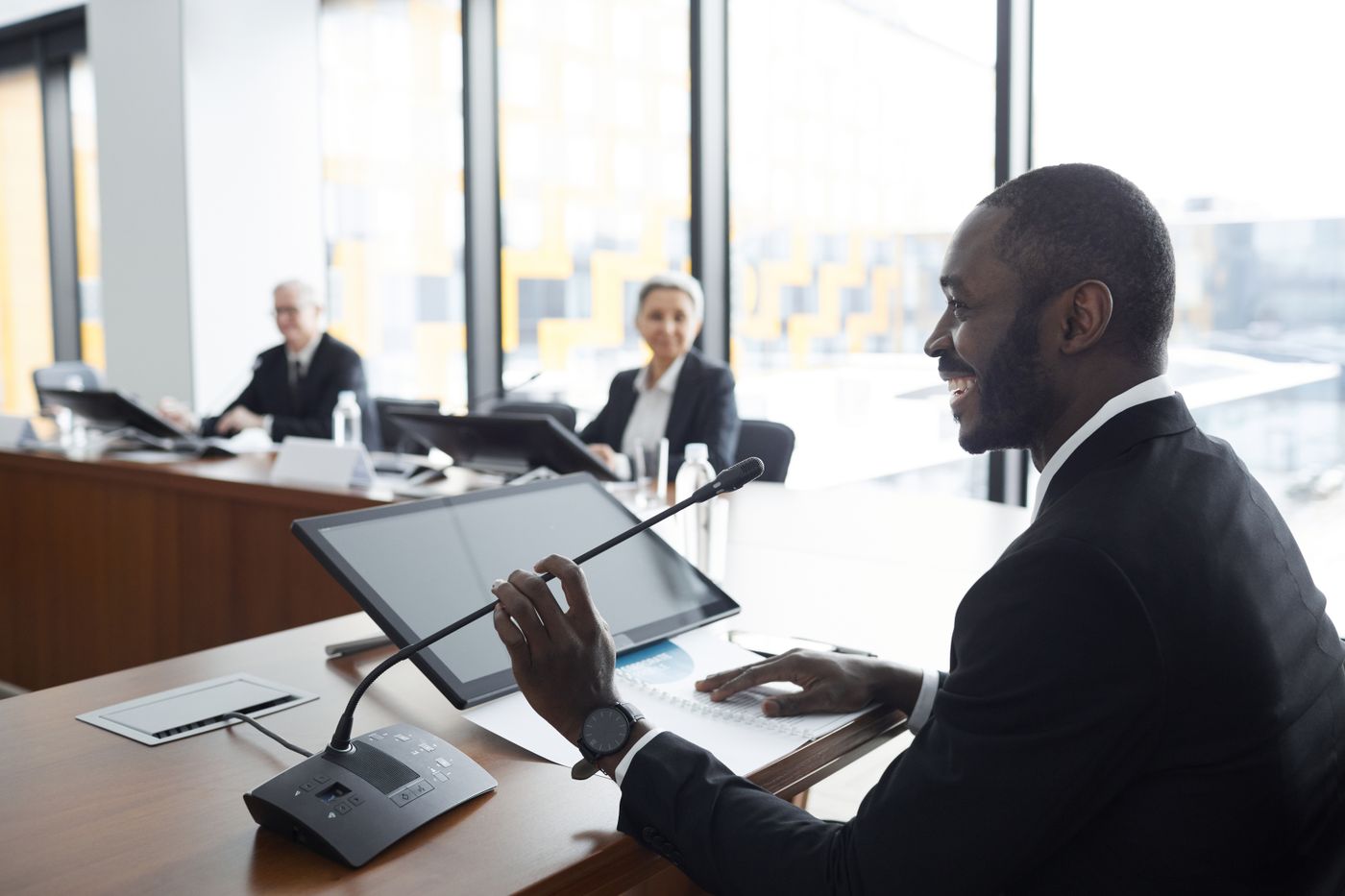 Smiling man in a dark suit sitting at a long conference table and speaking into a desk microphone beside a tablet, while two colleagues listen from the far end of the room by tall windows, a calm and ordinary turn to speak once the alarm has been switched off