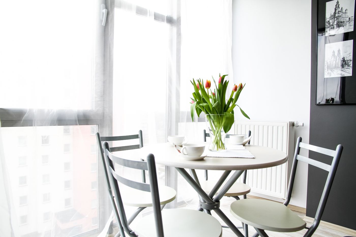 Small round white table by a bright curtained window, set with three white cups and saucers and a glass vase of red and yellow tulips, surrounded by empty grey chairs, the kind of laid place at the table that grief habits keep setting for someone who has gone
