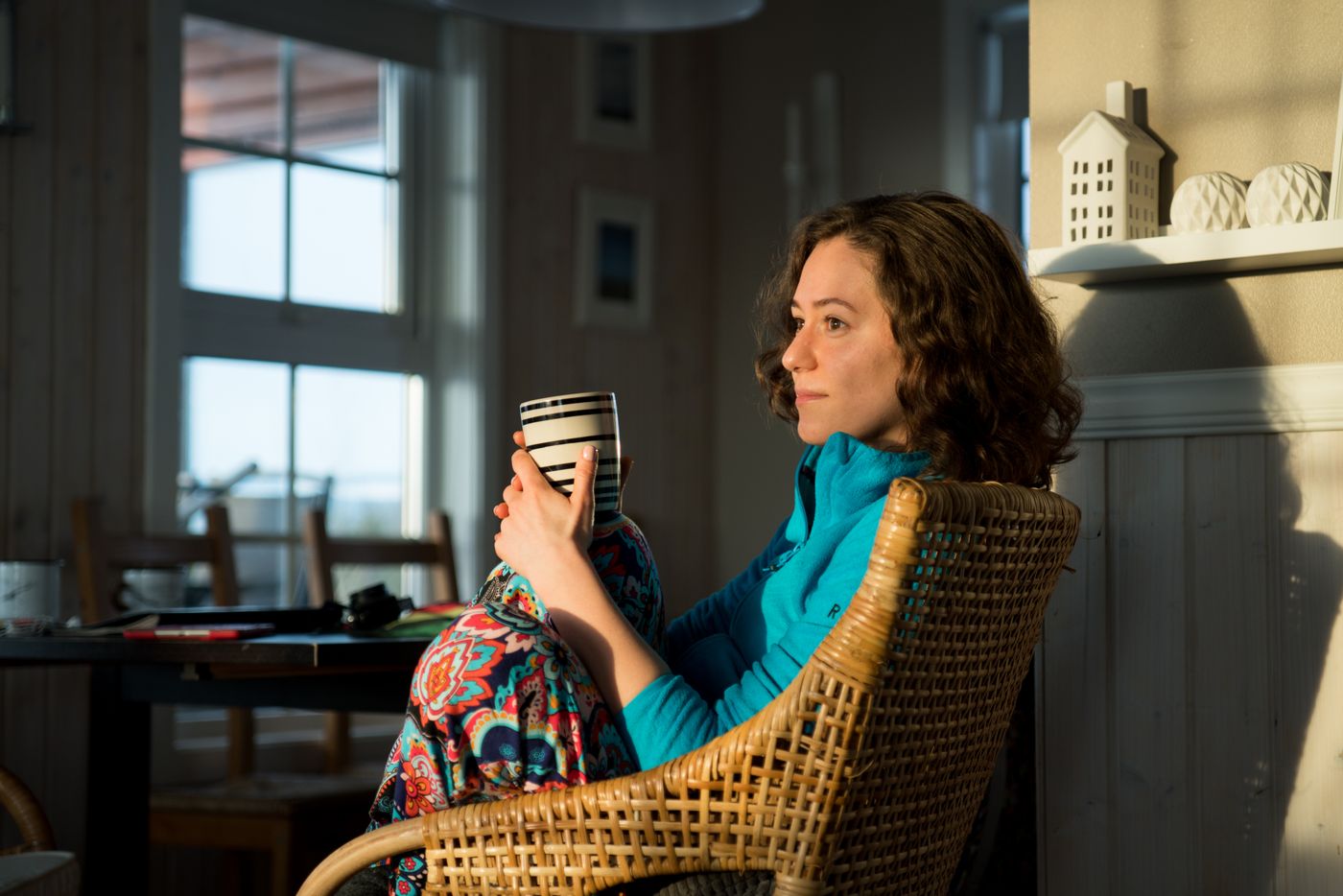 Woman with curly hair in a blue top relaxing in a wicker chair by a sunlit window, holding a striped mug in both hands and gazing calmly into the room, content in an ordinary moment without waiting for anything