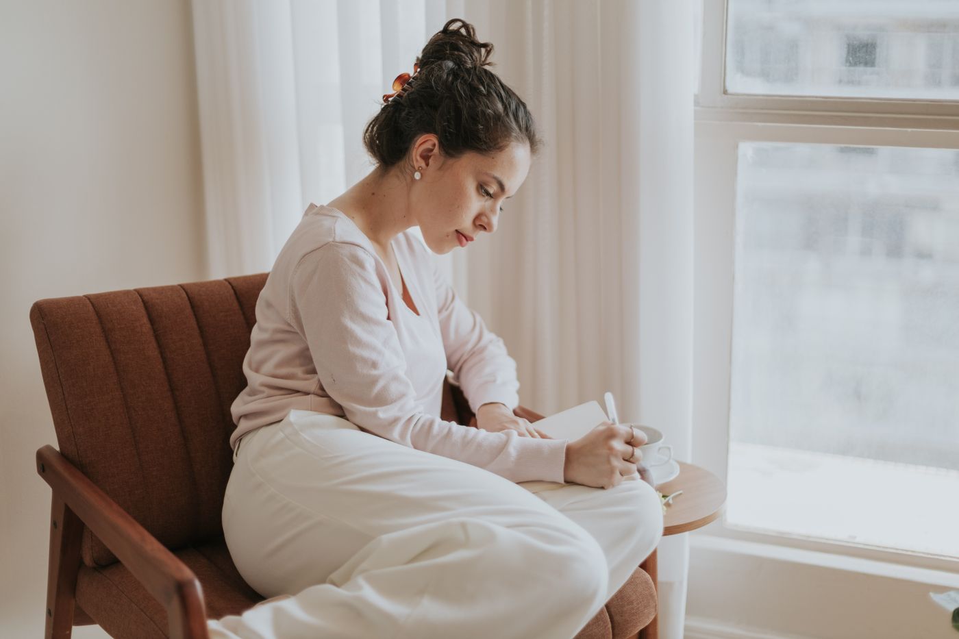 Woman in a pale jumper sitting in a brown armchair by a bright window writing in a notebook beside a cup of coffee, keeping a wobble log of the urge to sabotage a good week