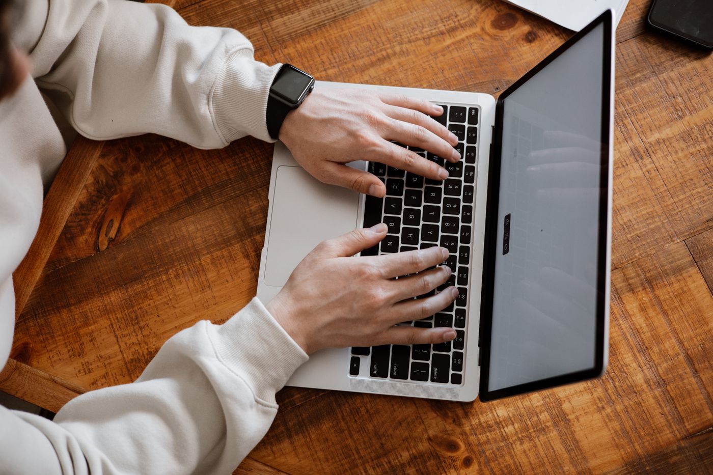 Overhead view of hands typing on a silver laptop at a wooden desk, the everyday moment when over-apologising slips into an email as sorry to bother you