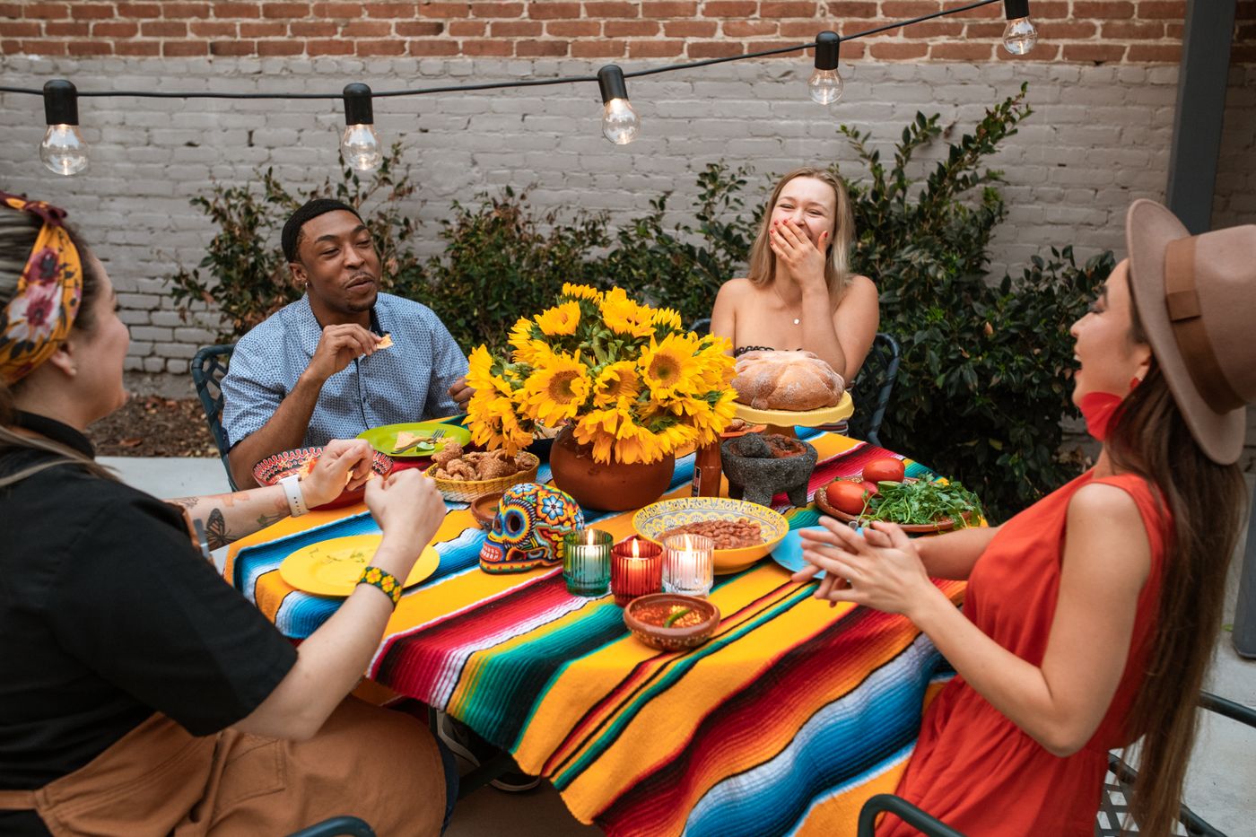 Four friends laughing around an outdoor table covered in a bright striped cloth, with sunflowers, candles and plates of food under festoon lights, the warm moments of an evening that the replay in your head conveniently leaves out