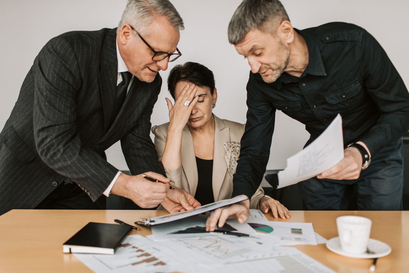 Stressed woman at a meeting table with her hand on her forehead while two male colleagues lean over her with papers and a pen, the pressure of other people's demands that makes saying yes feel like the only safe option