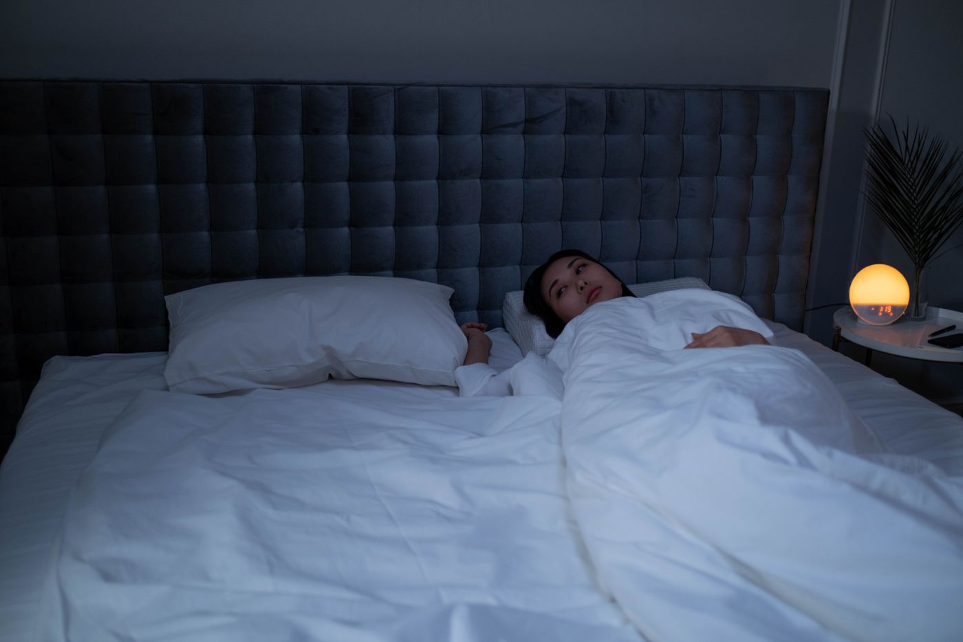 Woman lying awake under crisp white bedding in a stylish, dimly lit bedroom beside a glowing bedside lamp, eyes open in the small hours, the 3am audit when a life that looks fine on paper suddenly feels pointless