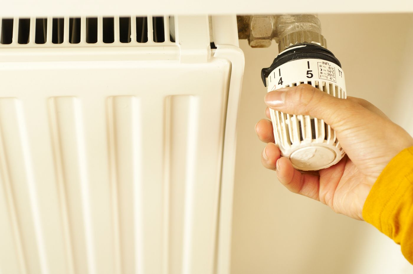 Hand in a mustard yellow sleeve turning the thermostatic valve on a white radiator, like the internal set point that switches the heat off when life gets warmer than you are used to