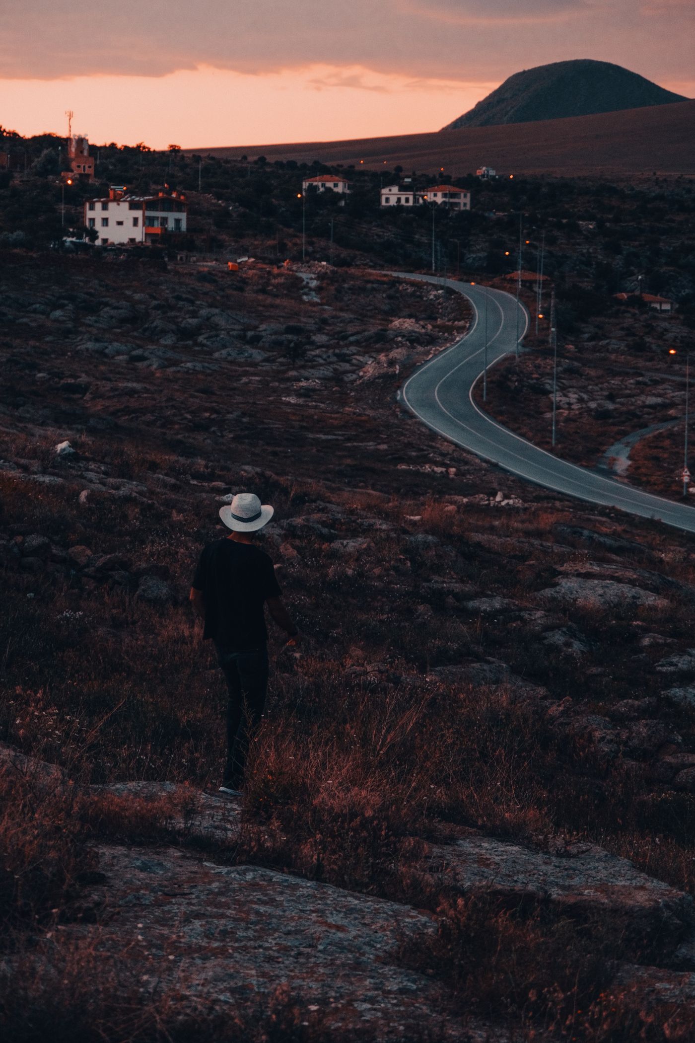 Man in a white hat standing alone on a rocky hillside at dusk, looking down a long winding road that curves away toward distant houses and a hill on the horizon, like a finish line that keeps moving further away