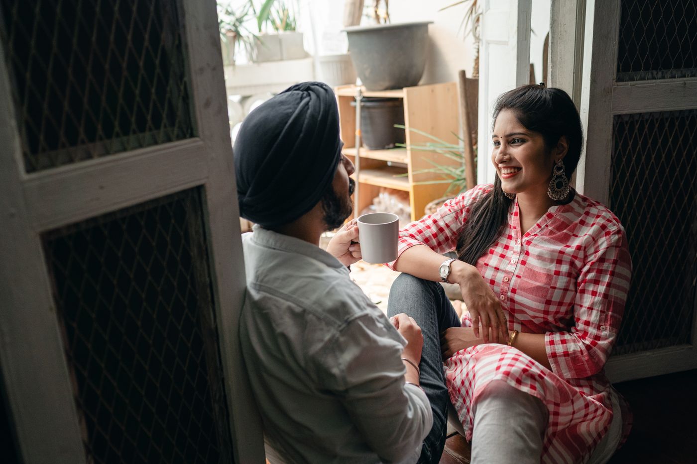 Smiling woman in a red check top sitting close to her partner in a sunlit doorway, the two of them talking face to face over mugs of tea, the easy conversation that replaces the silent treatment once the hidden sentence is said out loud