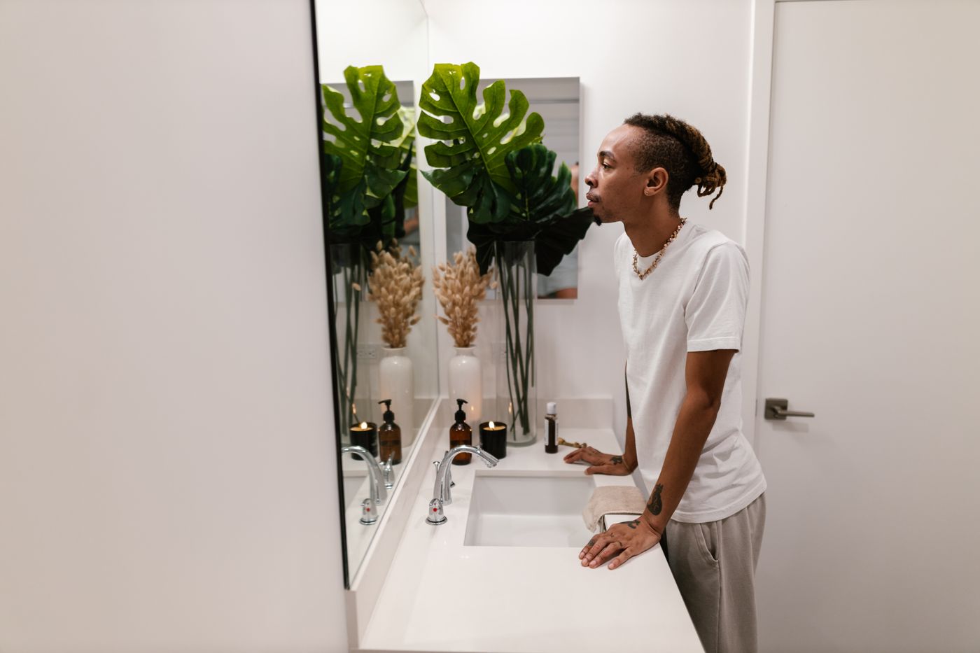 Man in a white T-shirt leaning on a bathroom sink and studying his reflection in the mirror, the everyday moment when negative self-talk starts its morning commentary