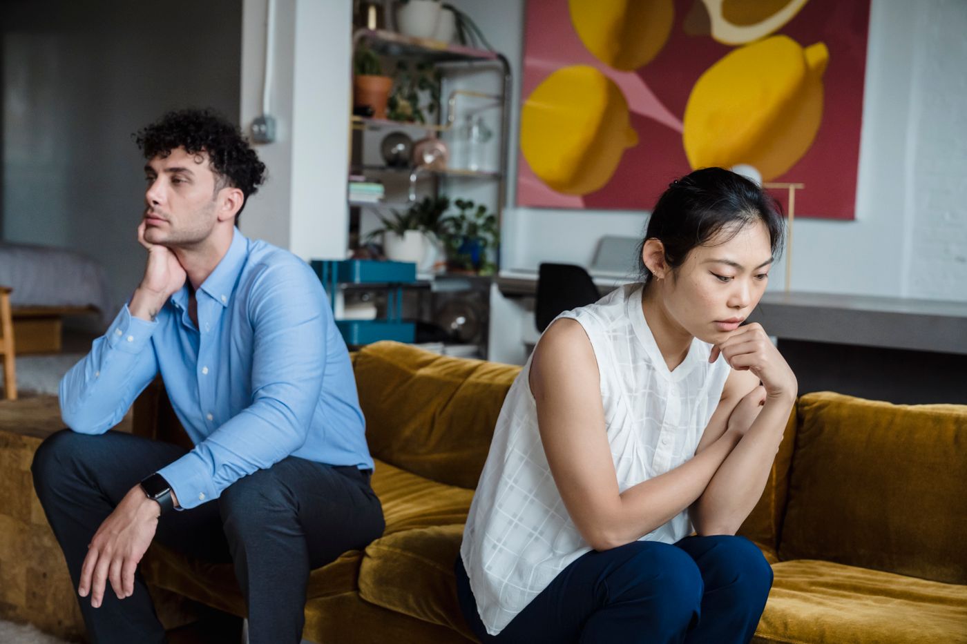 Young couple sitting apart at opposite ends of a mustard velvet sofa, the man staring off to one side with his chin in his hand and the woman looking down at the floor, the frozen standoff of the silent treatment after a small slight