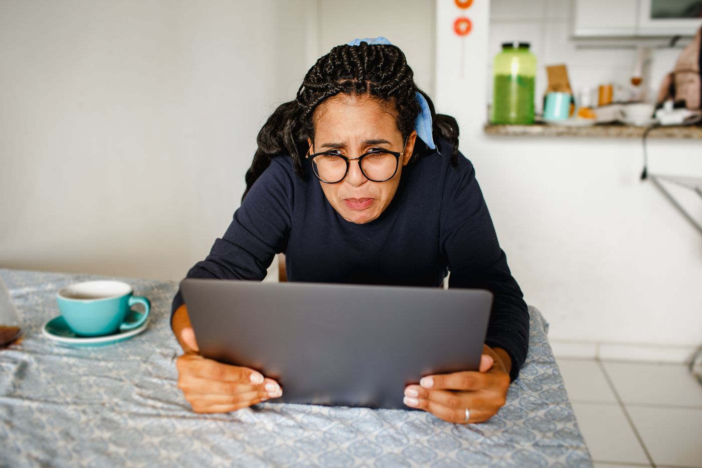 Woman in glasses leaning over a laptop at a kitchen table with a cup of coffee beside her, frowning at the screen on what should be a quiet day off, pulled back to work by the low hum of rest guilt
