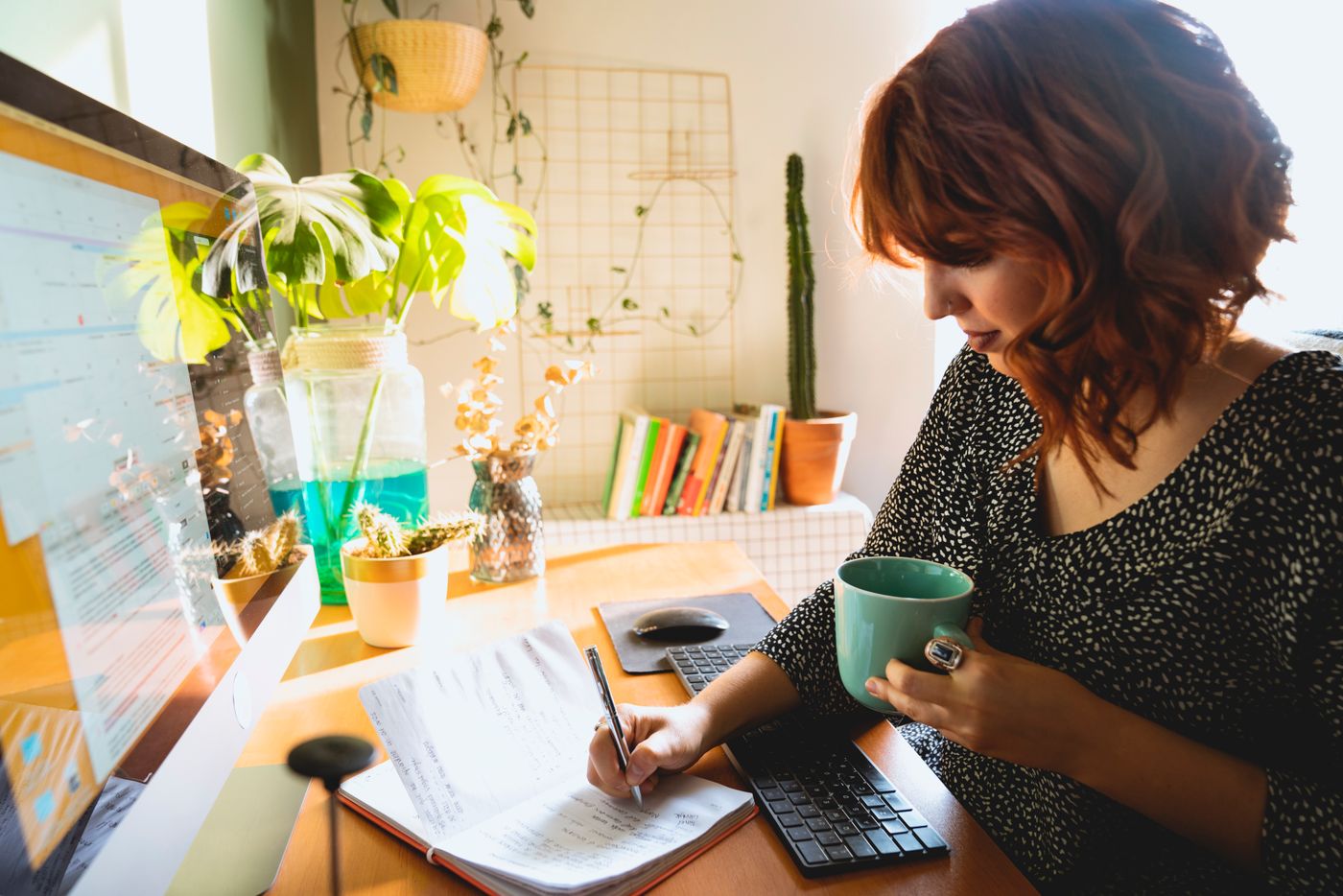 Woman writing notes in an open notebook at a sunny home desk while holding a green mug, the simple act of putting the week's worries on paper so Sunday dread gets an appointment instead of running the whole evening