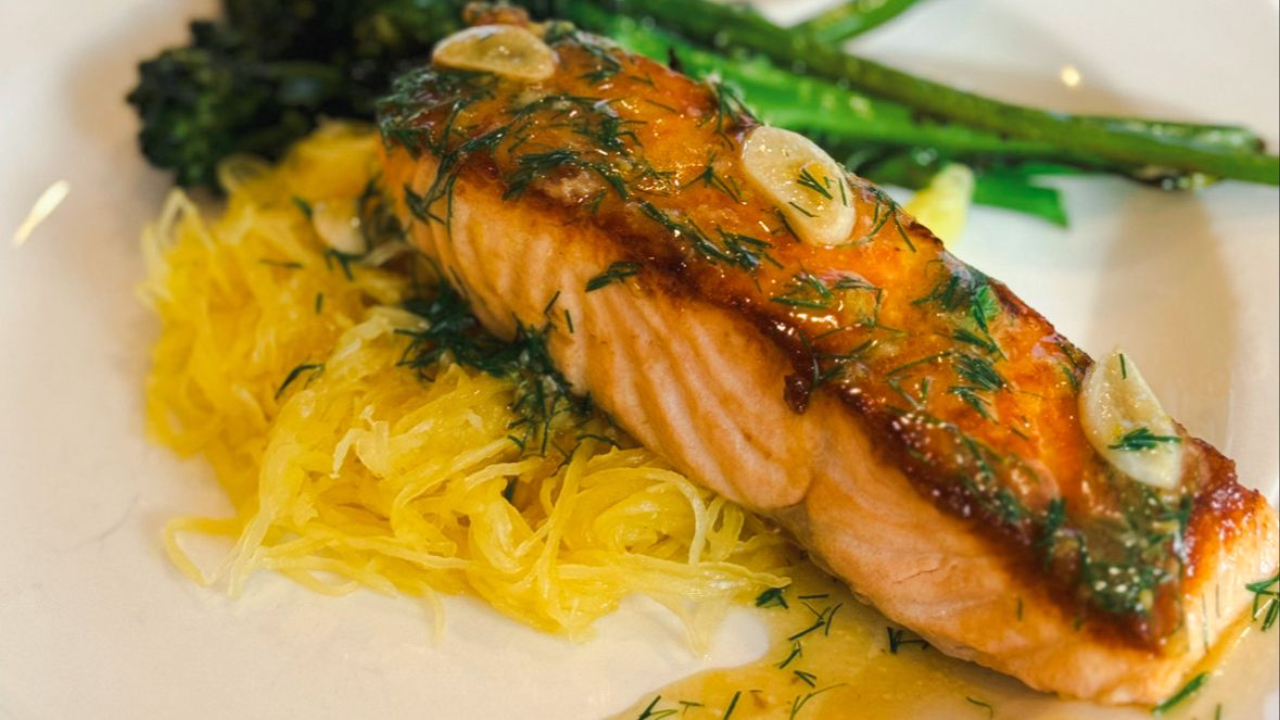 Grilled salmon fillet garnished with dill and garlic, served alongside spaghetti squash and asparagus on a white plate, presenting a healthy and flavorful meal option.