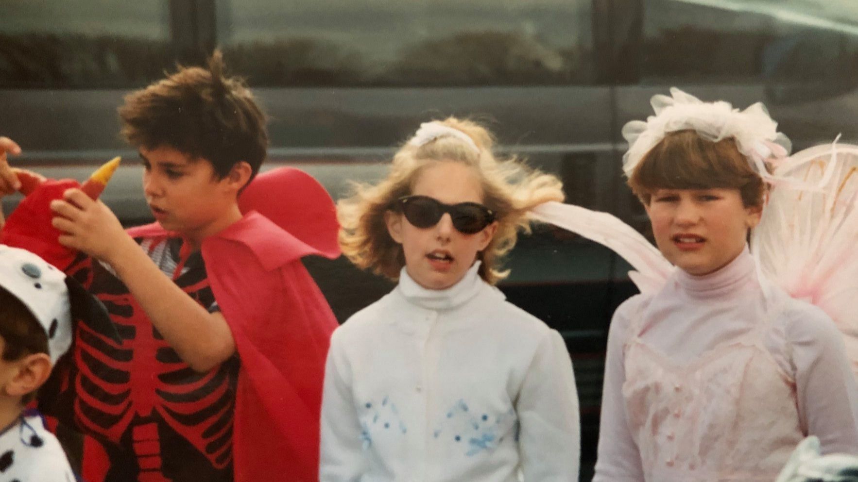 Three children in Halloween costumes enjoying a festive celebration outdoors, with a boy in a red devil costume adjusting his mask, a girl in the middle channeling a pop star look with sunglasses, and another girl dressed as an angel with white wings and a headband.