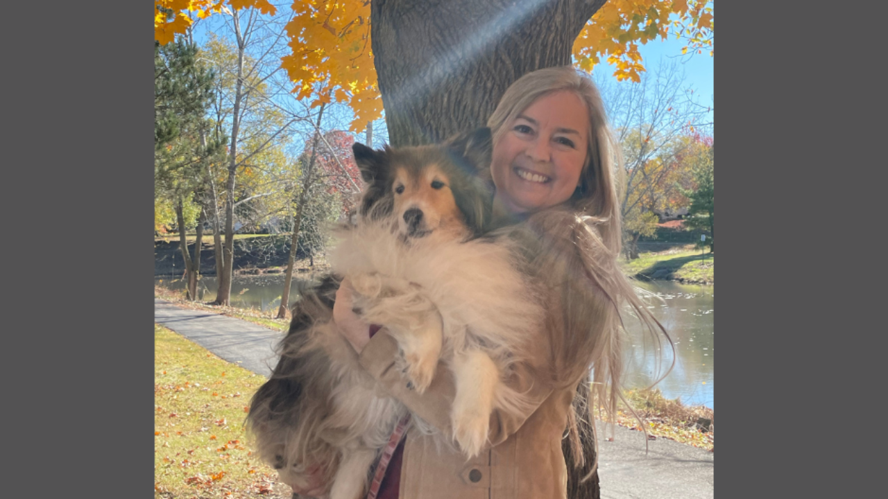smiling woman holding a sheltie dog on a sunny fall day