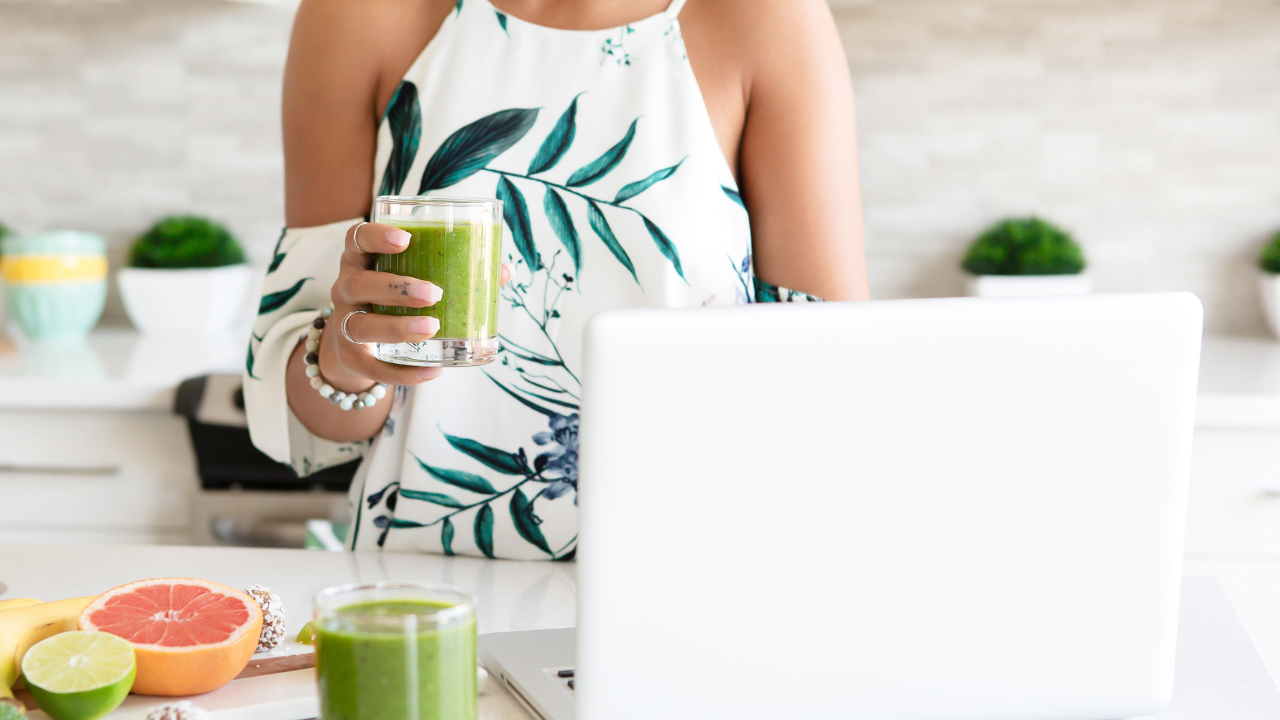 Woman making a green smoothie while using a laptop, representing the struggle between healthy routines and a busy mind
