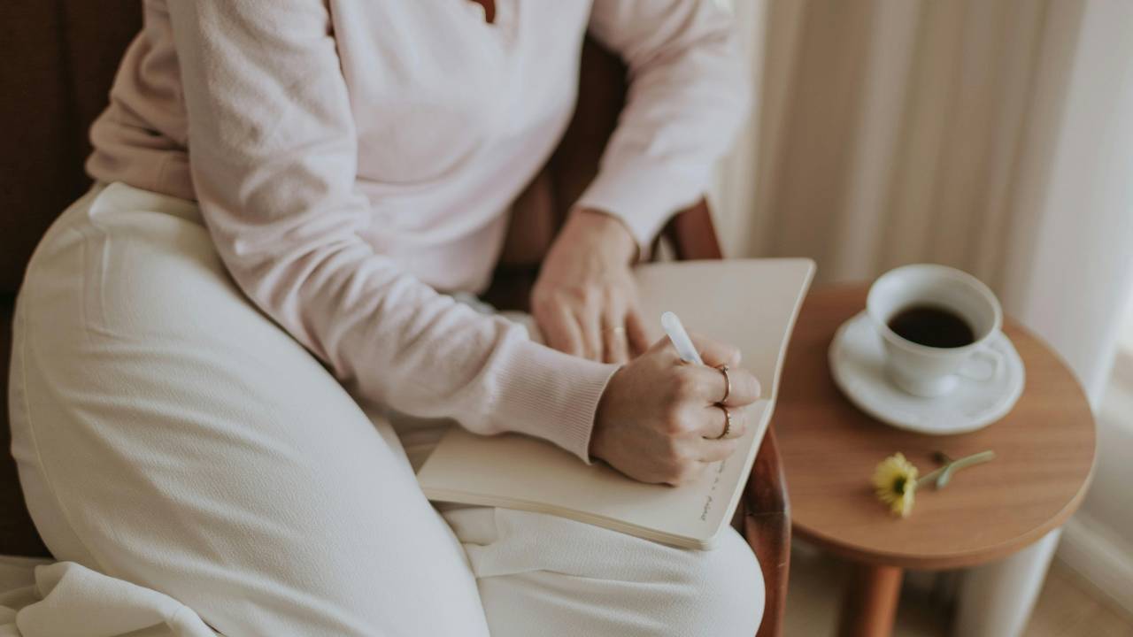 A person dressed in all white clothes sitting on the couch comfortably, writing in their notebook, with a coffee on the side table.