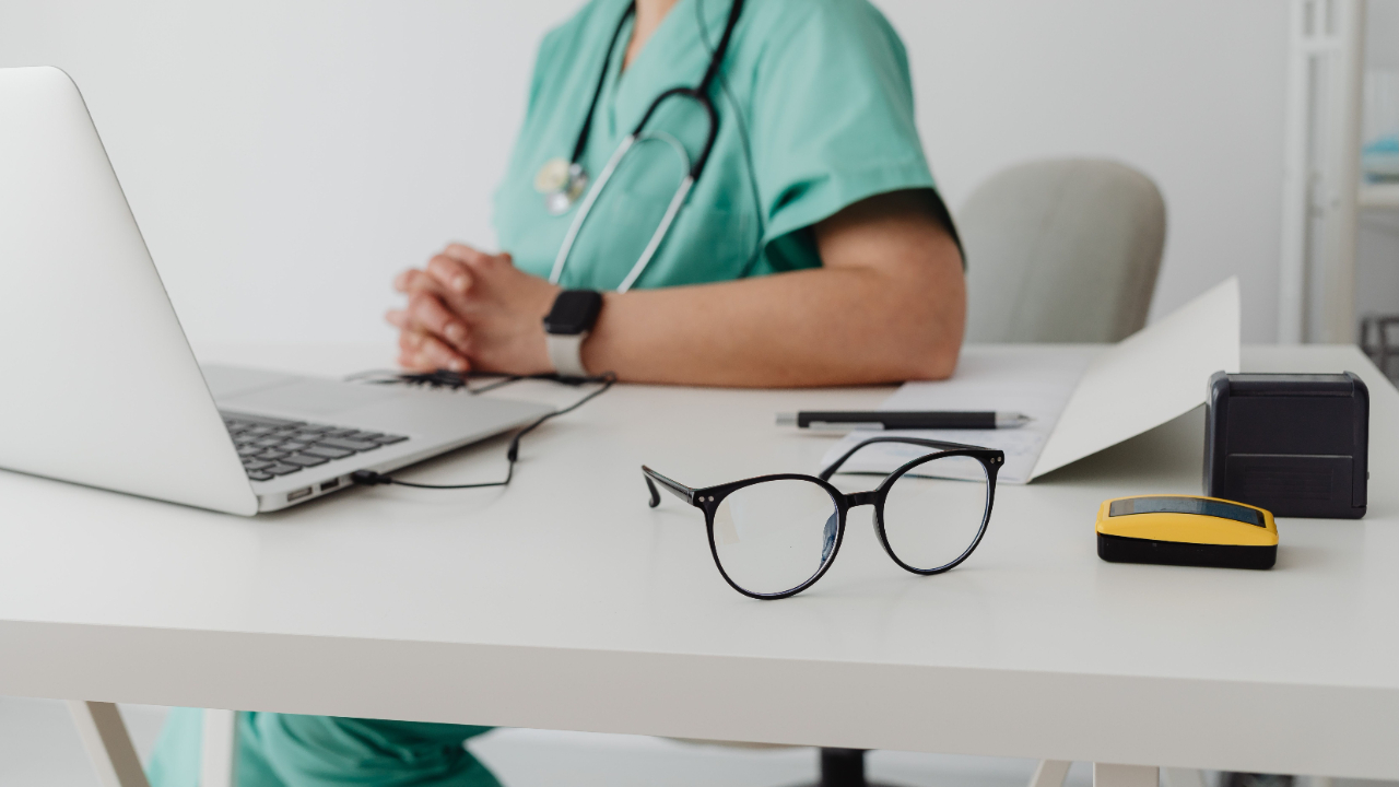 Image of a doctor sitting behind a desk with a computed and eyeglass at the table