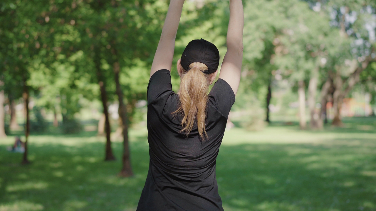 A woman stretches in the morning sunlight surrounded by green trees, symbolising energy restoration, flow, and mindful movement