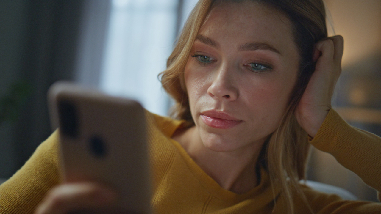 Woman in a mustard sweater looking thoughtfully at her phone, illustrating feelings of self-doubt and comparison while scrolling social media.