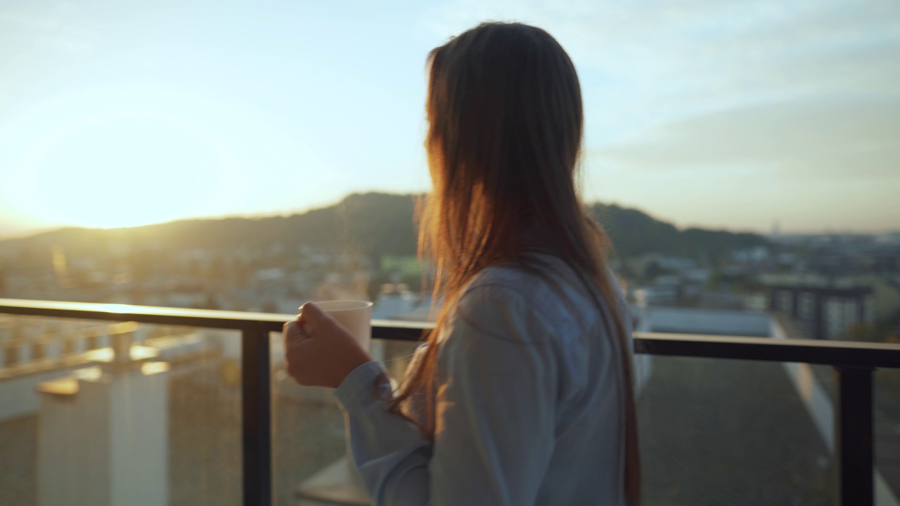 A woman stands on a balcony at sunrise holding a cup of coffee, reflecting calmly and regaining mental clarity and energy