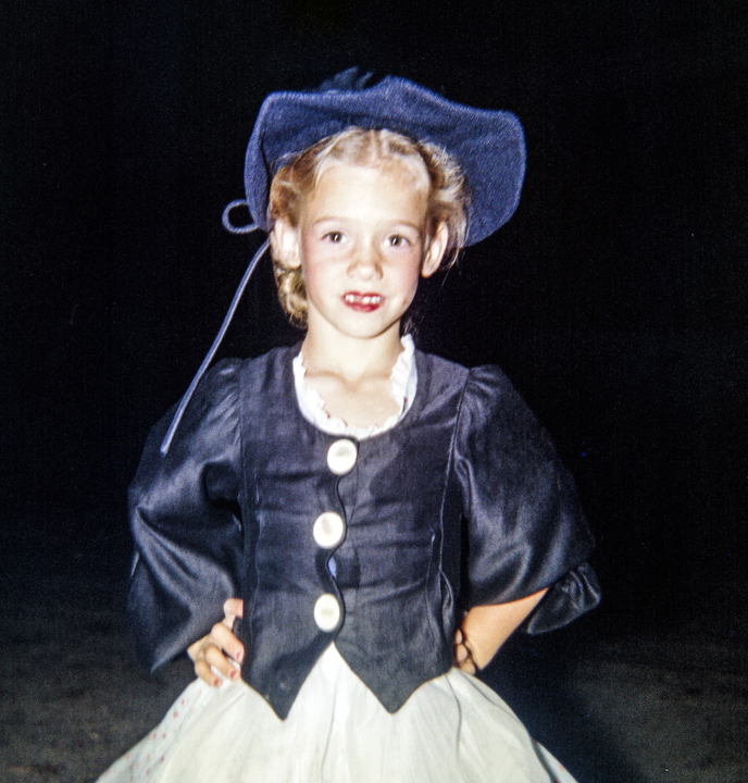 Young girl in a blue hat and dark vest with her hands on her hips.