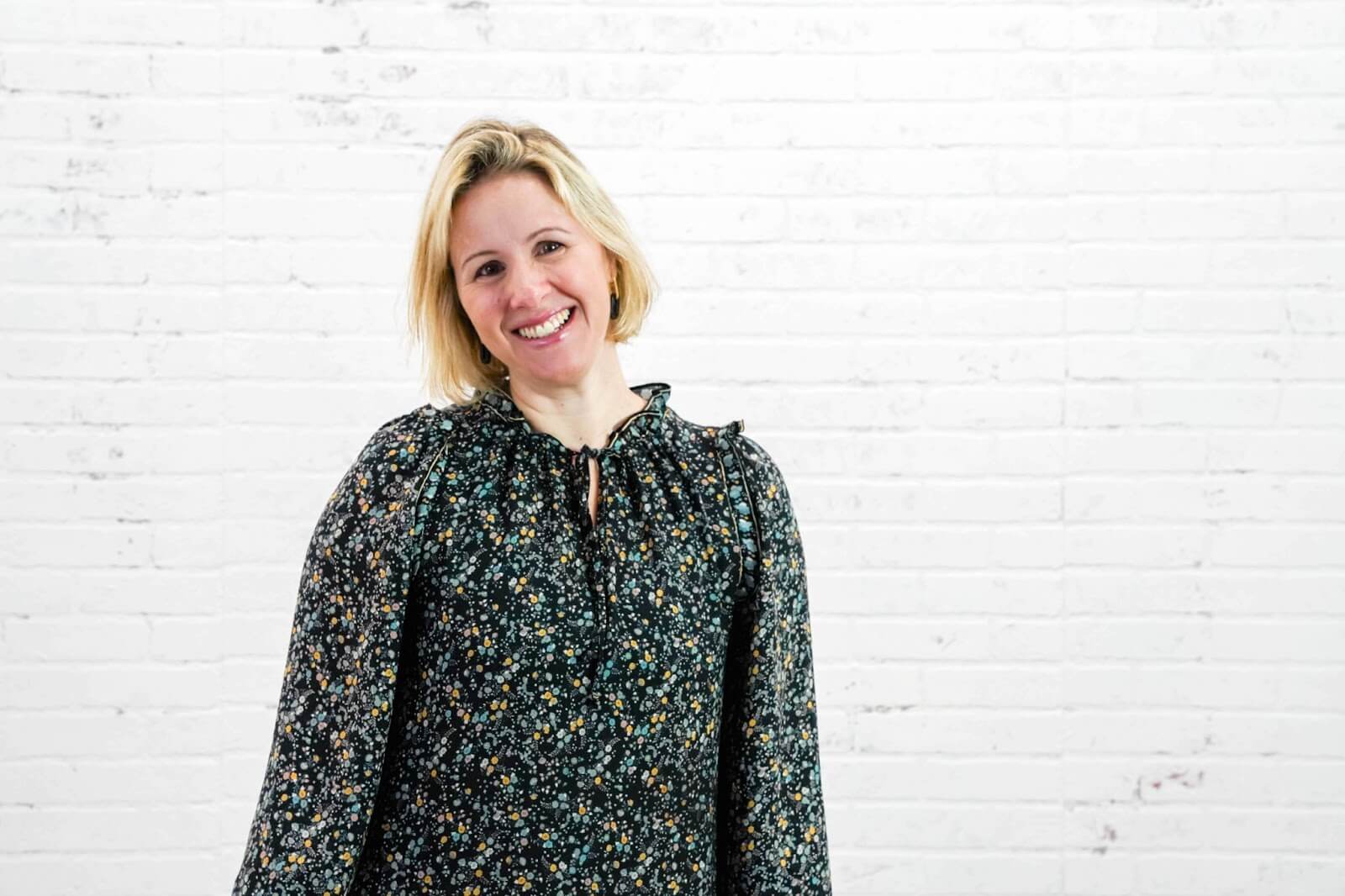 successful home stager in a black floral dress stands against a white brick wall