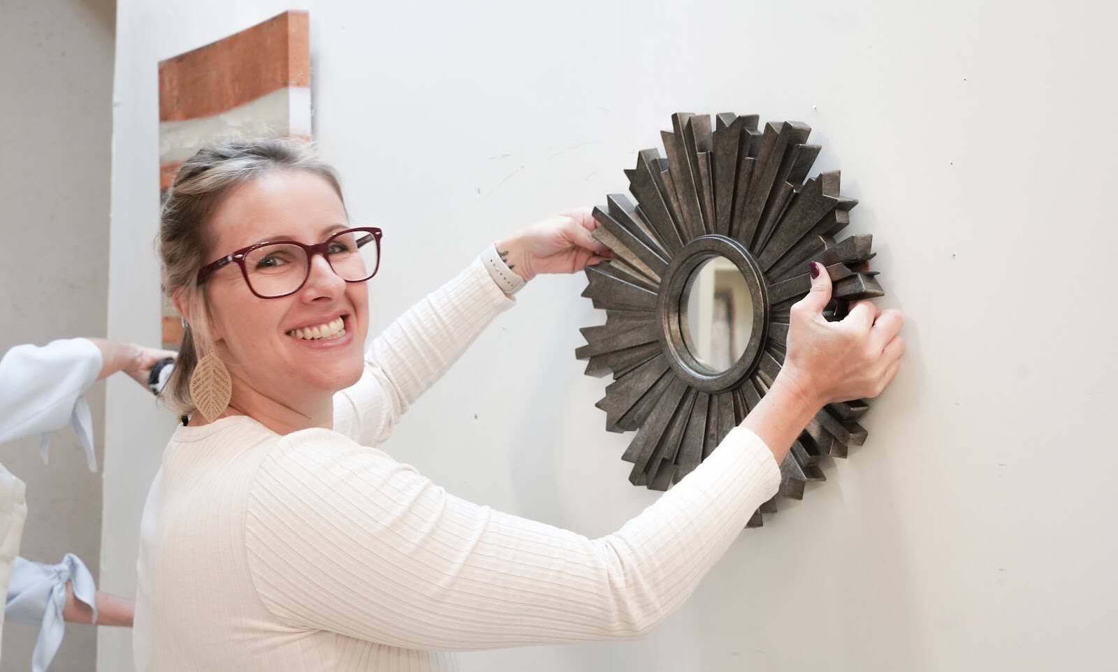 Photo of a blonde white woman with glasses hanging a round mirror on the wall at the Staging Studio Master Class