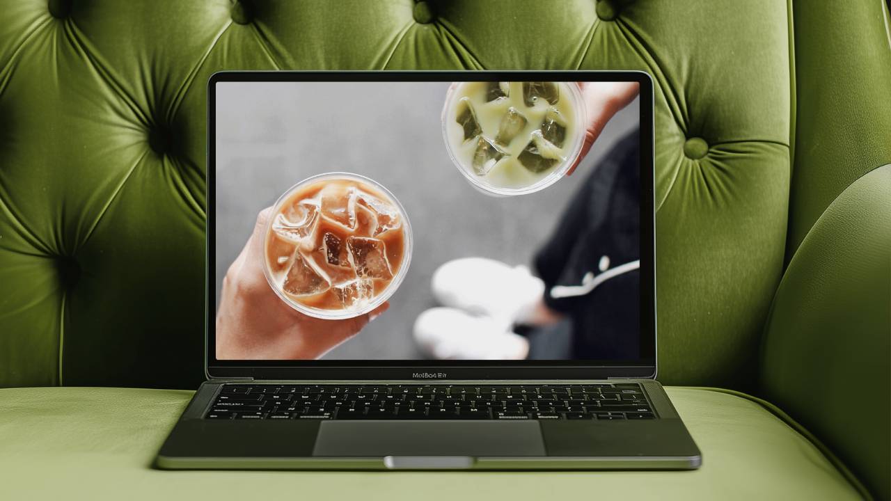 A laptop sits on a green tufted couch, displaying a close-up image of two special education teachers holding iced coffee drinks — one creamy brown and one matcha green. The scene feels relaxed and modern, suggesting remote work, collaboration, and creative entrepreneurship.