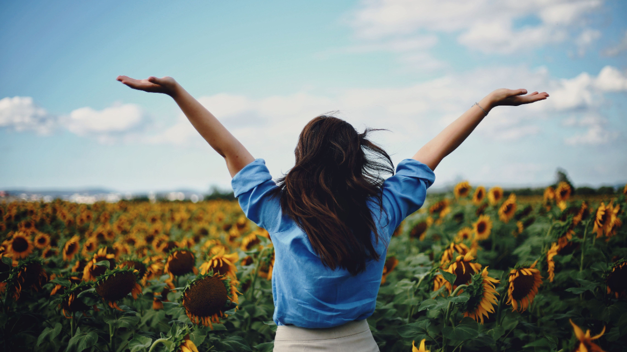Person standing with arms raised open in a sunflower field under a partly cloudy sky.