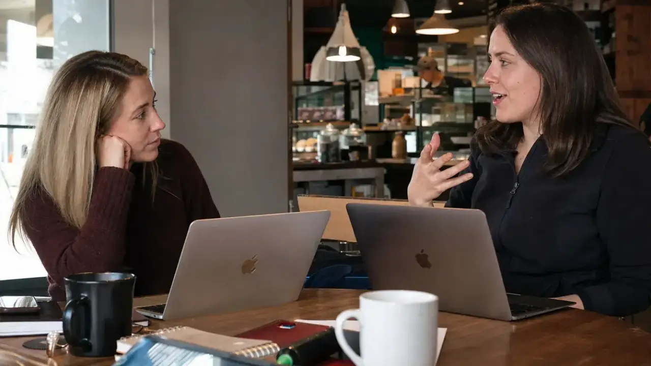 Dos profesionales reales conversando en una cafetería con portátiles abiertos entre ellas, luz natural de día, ambiente de trabajo colaborativo real