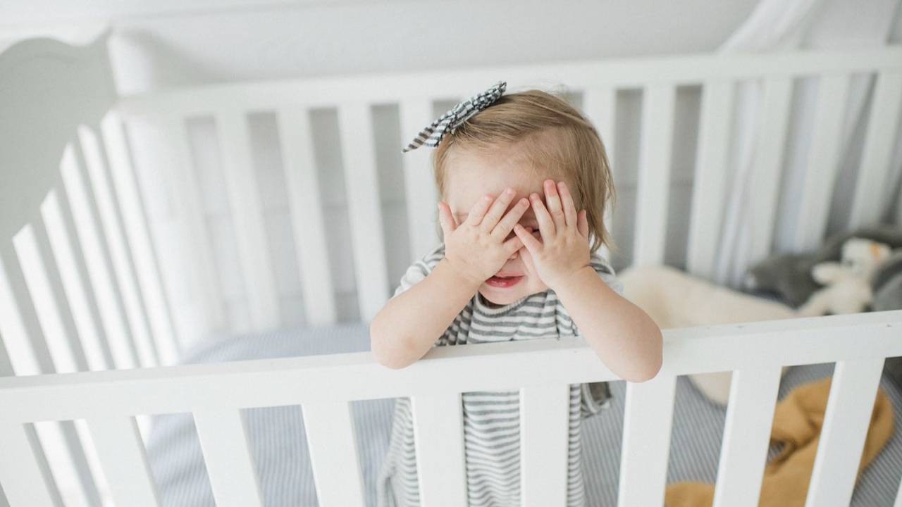 A baby standing up awake in their crib covering her eyes